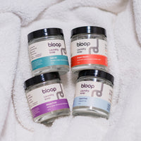 Four jars of Bloop laundry soap, featuring scents like Salt Air and Super Bloom, sit on a soft white towel.
