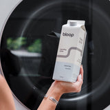 A hand holding a carton of Bloop unscented laundry soap displays it in front of a washing machine.
