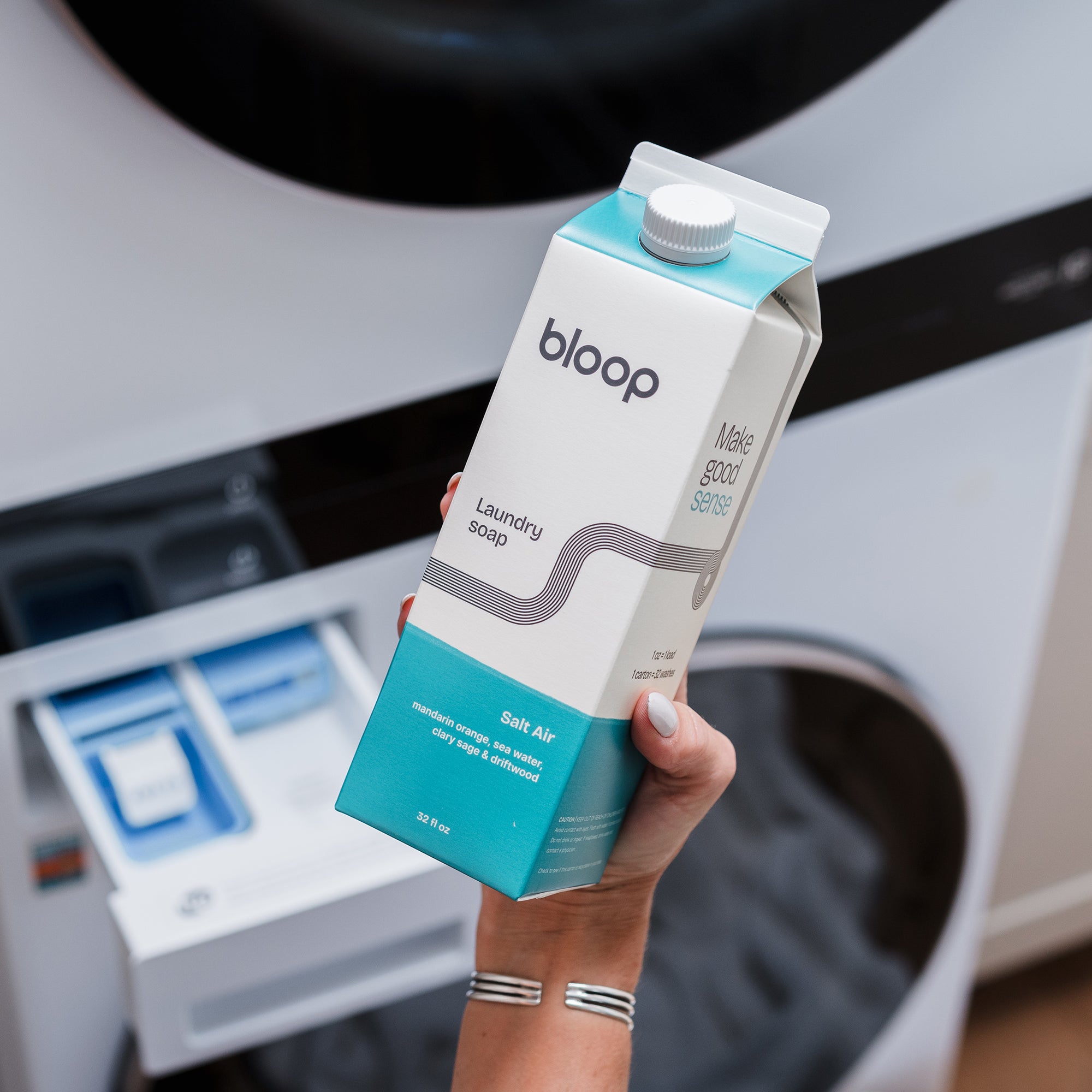A woman's hand holds a carton of Bloop laundry soap, Salt Air scent, near an open washing machine detergent drawer.
