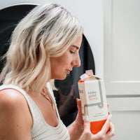 A woman smells the fresh scent of Bloop laundry soap in its eco-friendly carton, standing next to her washing machine.
