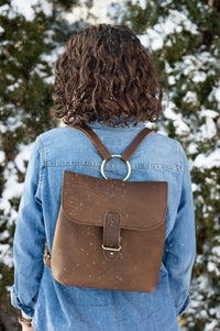 A woman wears a stylish brown leather backpack with gold ring detail and a denim jacket while standing outdoors in a gentle snowfall.
