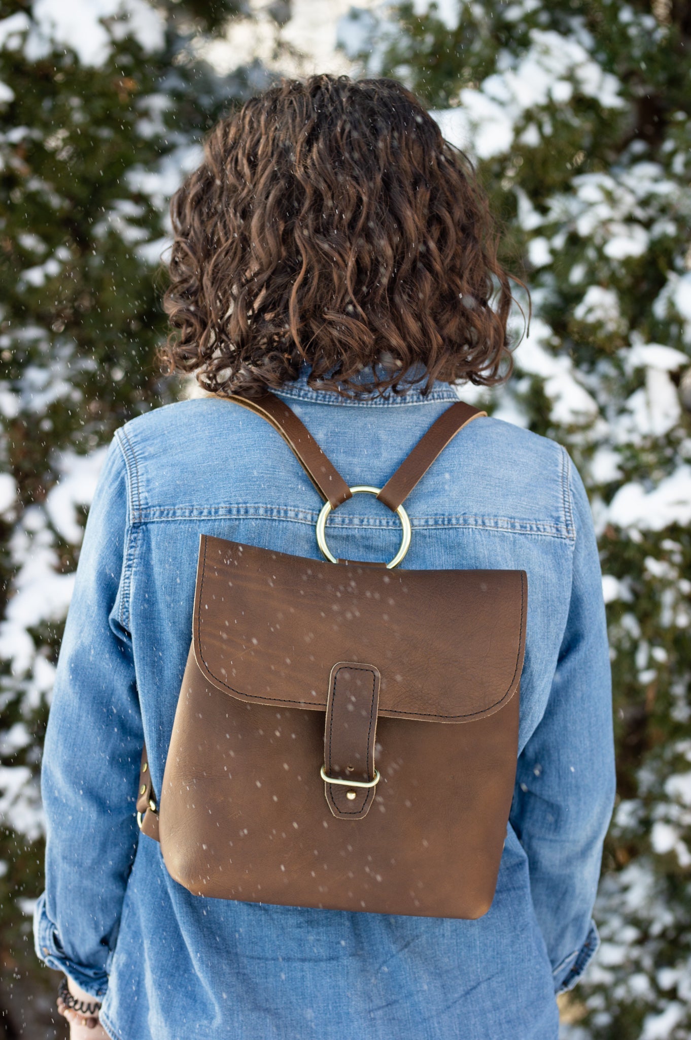 A woman wears a stylish brown leather backpack with gold ring detail and a denim jacket while standing outdoors in a gentle snowfall.
