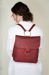 A woman models a stylish red leather backpack with gold hardware and unique circle detail.
