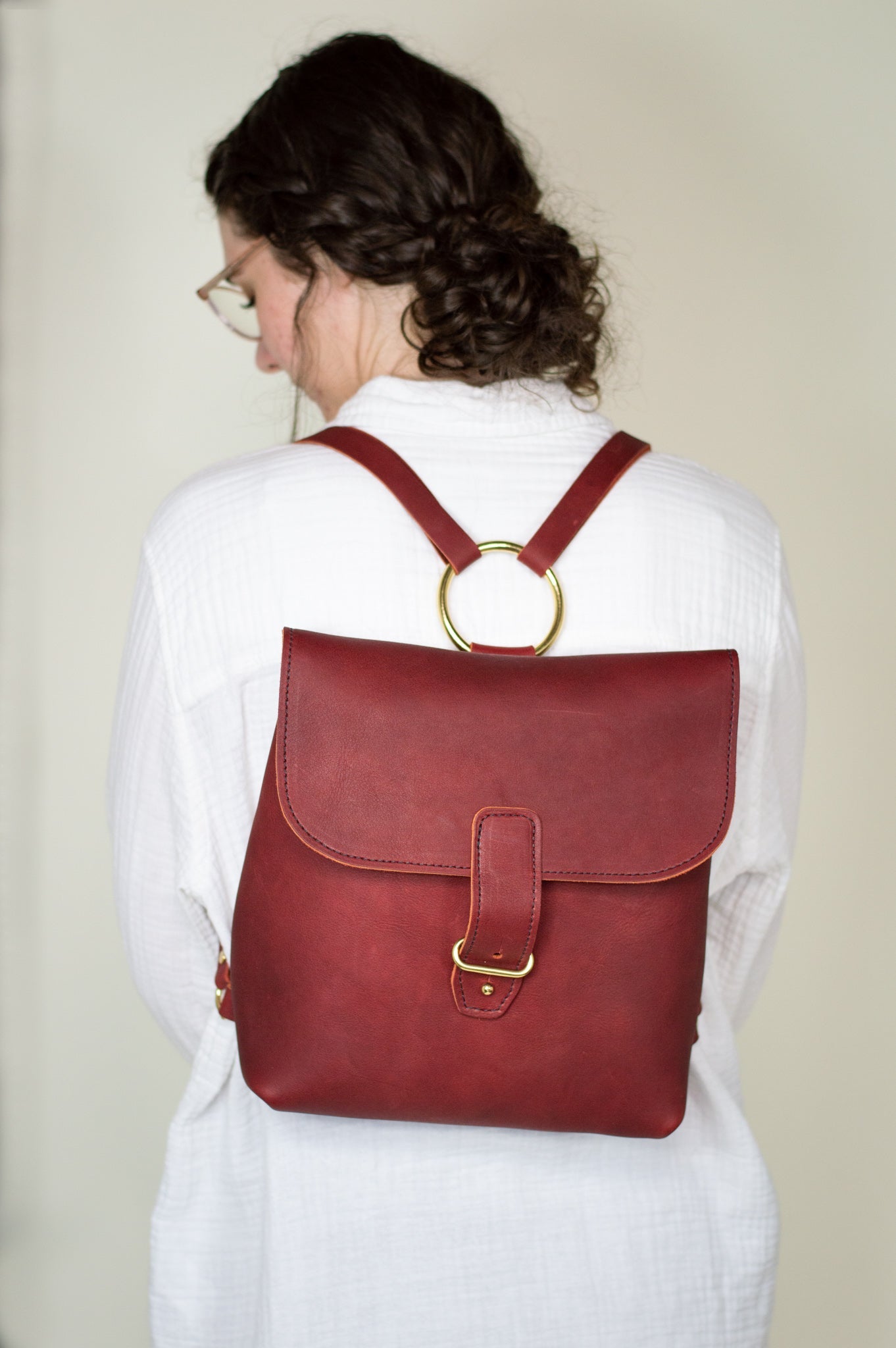 A woman models a stylish red leather backpack with gold hardware and unique circle detail.

