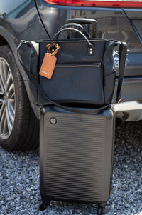 Black carry-on suitcase sits beneath a leather travel tote bag near a parked car.
