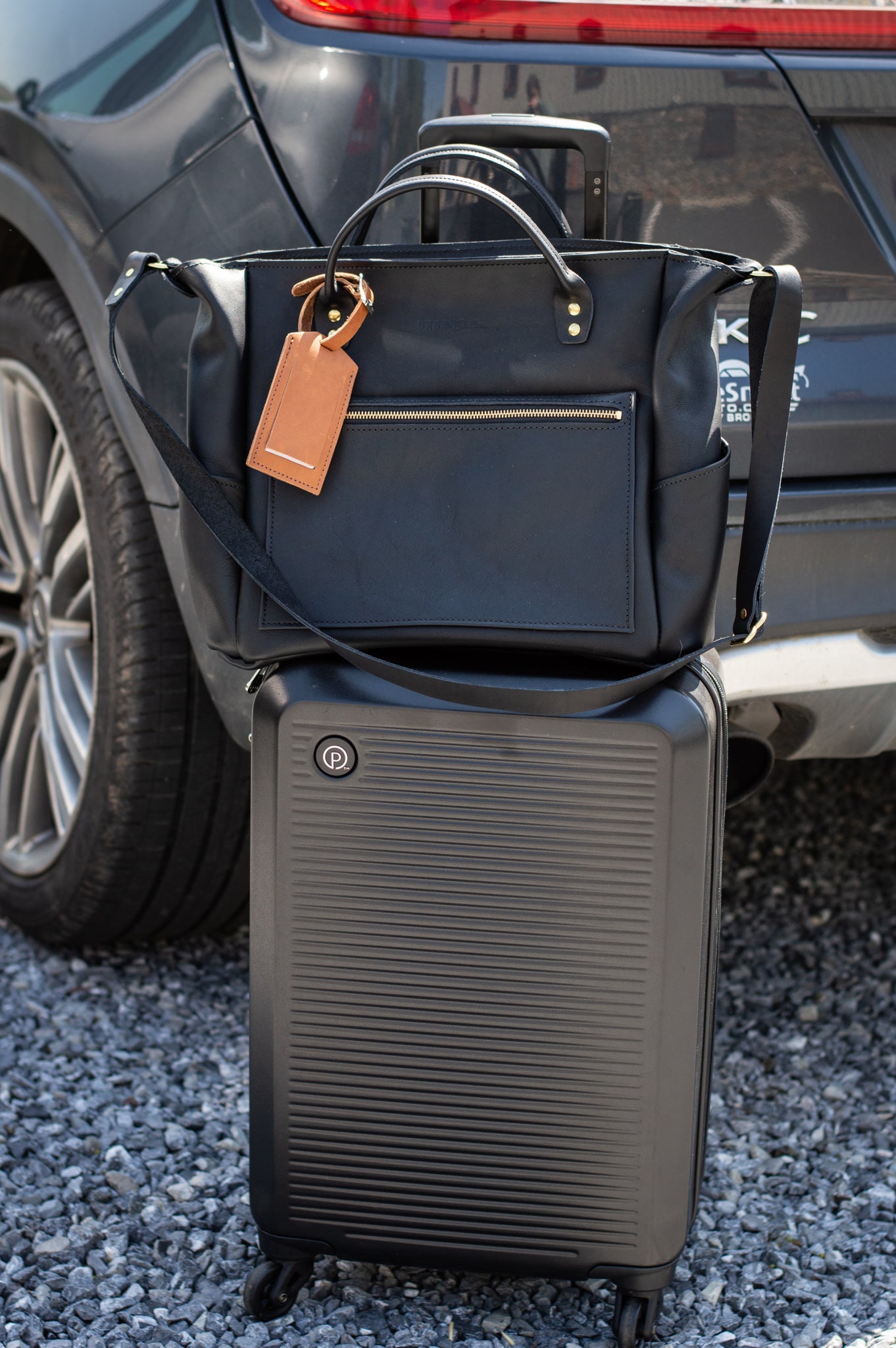 Black carry-on suitcase sits beneath a leather travel tote bag near a parked car.
