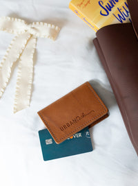Lifestyle flatlay features an Urban Southern leather wallet with a Discover credit card, a book, and a decorative ribbon on white sheets.
