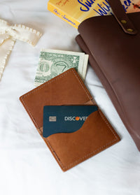 A minimalist brown leather wallet holds a Discover credit card and cash on a white bedsheet next to a summer reading book and a brown tote bag.
