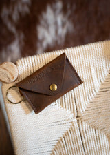 A brown leather Urban Southern wallet with a brass snap rests on a woven cream chair.
