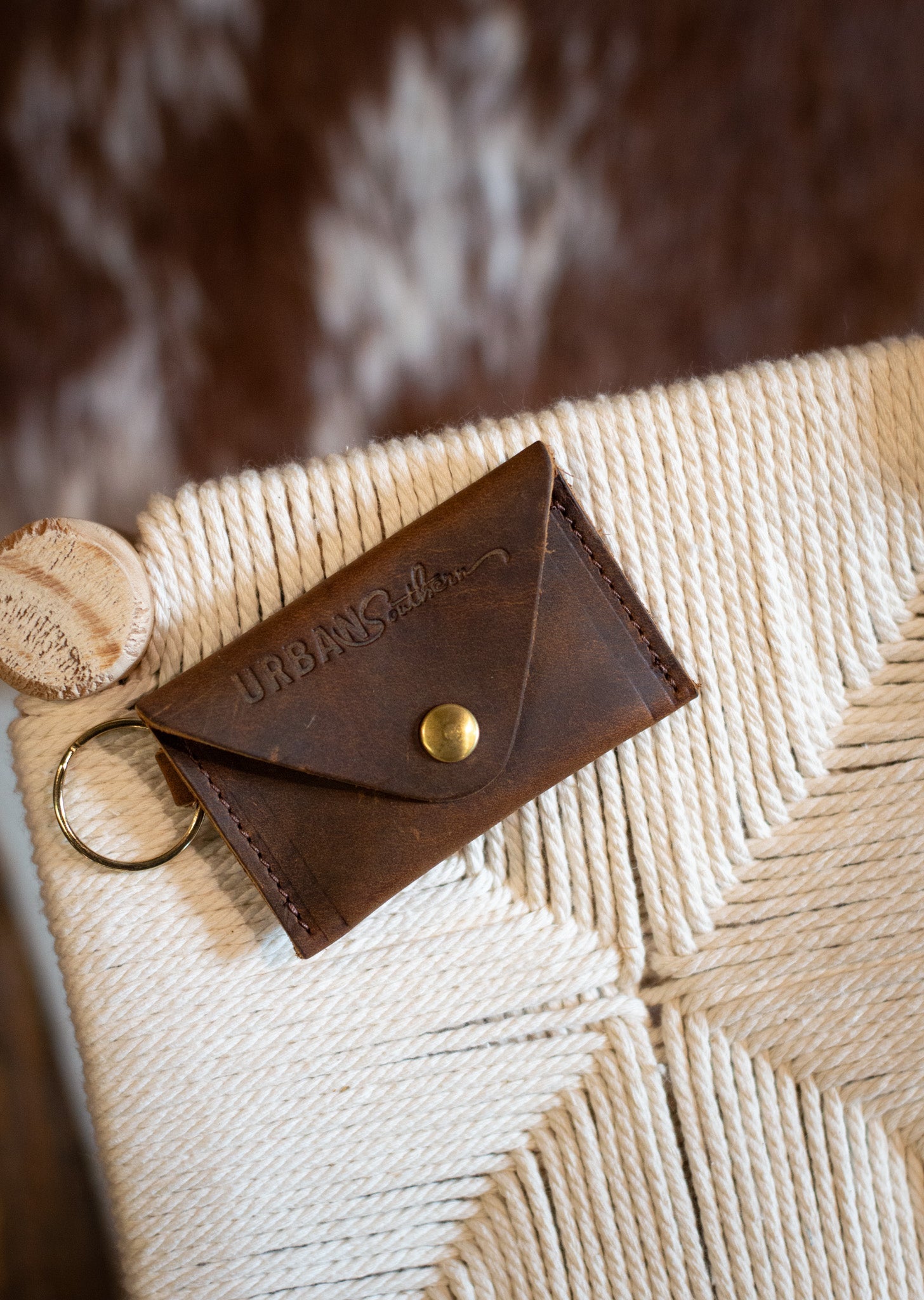 A brown leather Urban Southern wallet with a brass snap rests on a woven cream chair.
