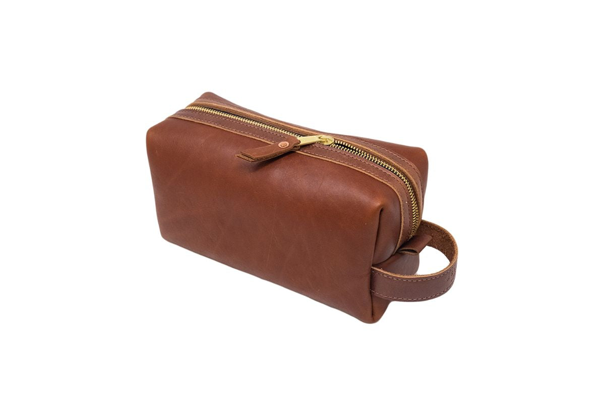 A brown leather dopp kit bag displays a golden zipper and a side handle, ready for travel.
