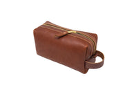 A brown leather dopp kit bag displays a golden zipper and a side handle, ready for travel.
