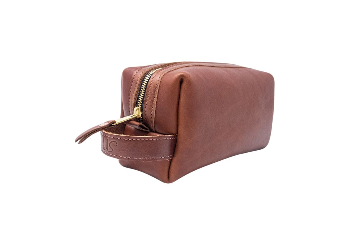 A brown leather toiletry bag with a gold zipper and stitched handle sits on a white background.
