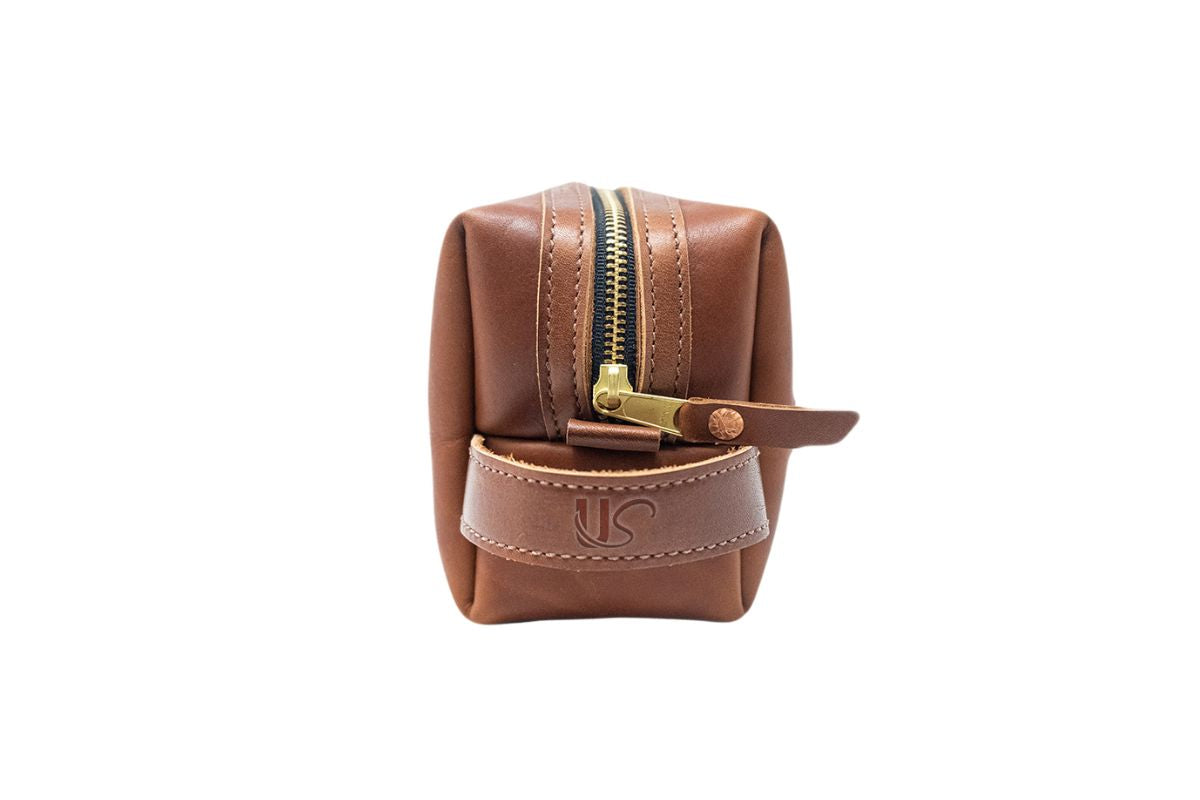 A brown leather Dopp kit displays a gold zipper and embossed "US" logo.
