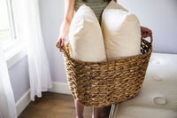 Person holding a wicker basket with two pillows inside, in a room setting.