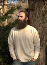 Bearded man with long hair leans against a tree, looking thoughtfully into the distance outdoors.
