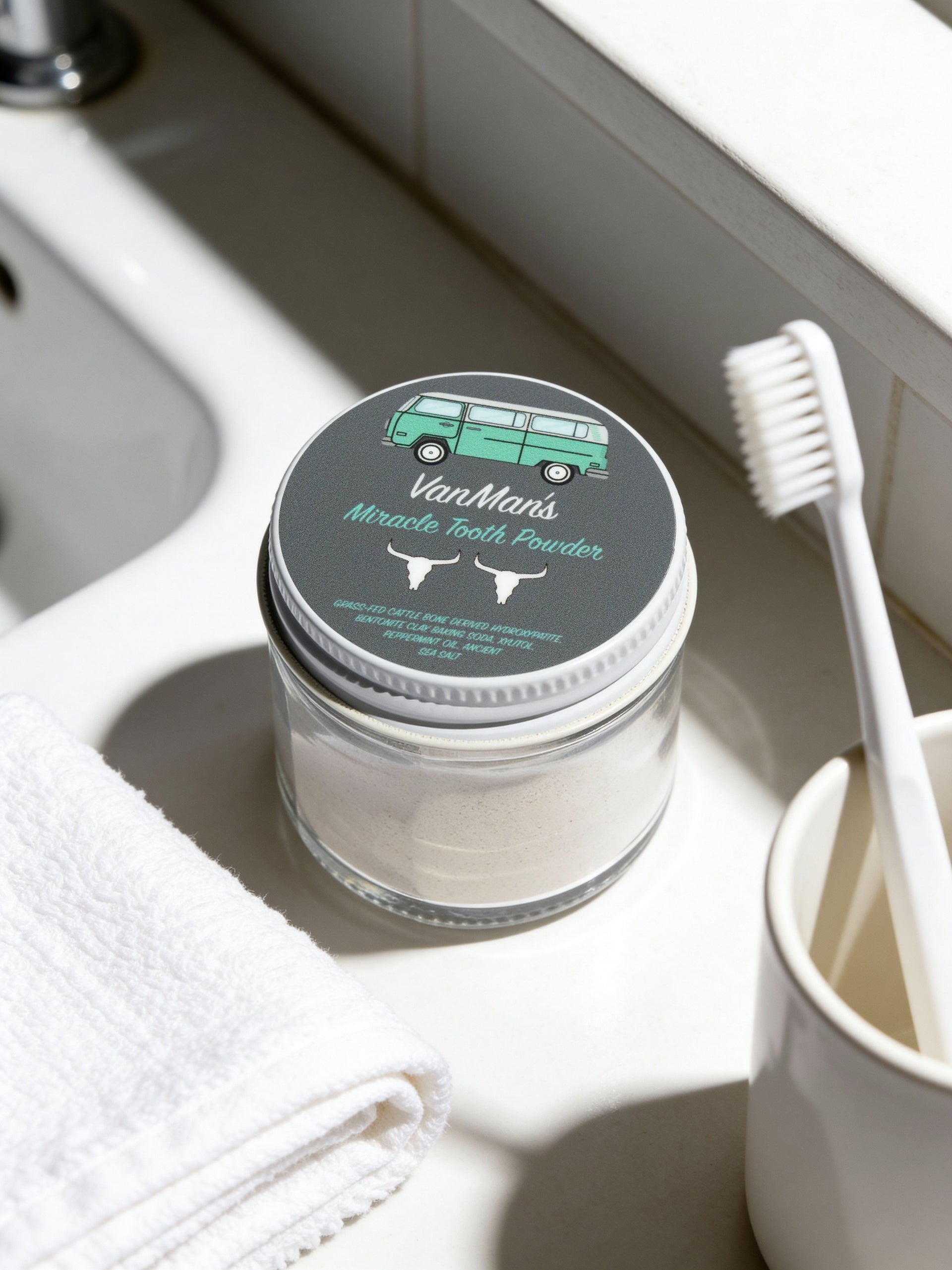 Jar of VanMan's Miracle Tooth Powder on a bathroom counter with a toothbrush and towel.