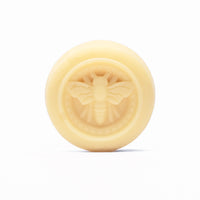 A round, pale yellow lotion bar displays a raised honeybee design.
