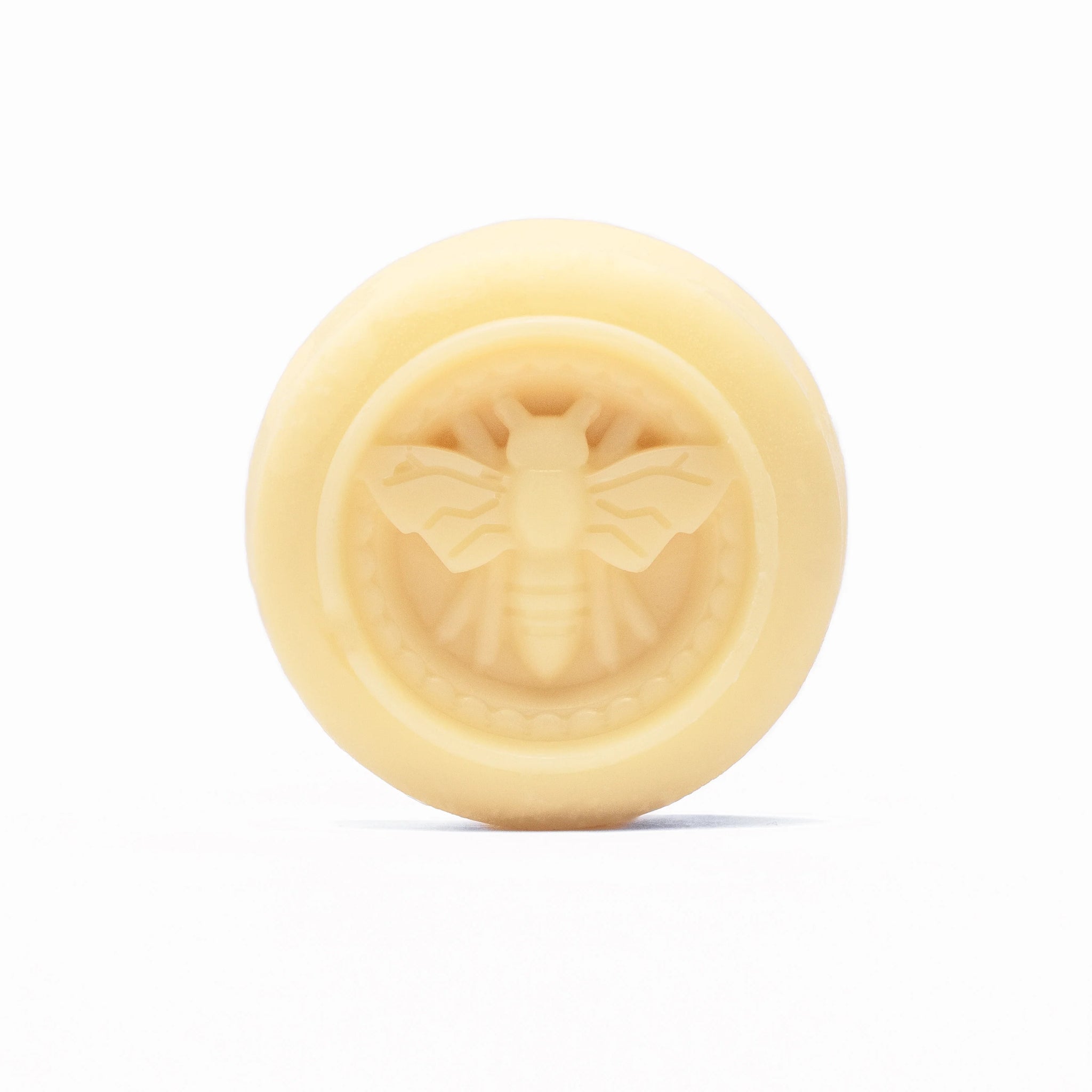 A round, pale yellow lotion bar displays a raised honeybee design.

