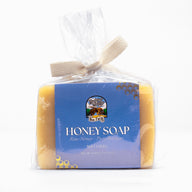 A bar of Register Family Bee Farm honey soap, made with raw honey and pure beeswax, sits wrapped in clear packaging with a woven ribbon tie.
