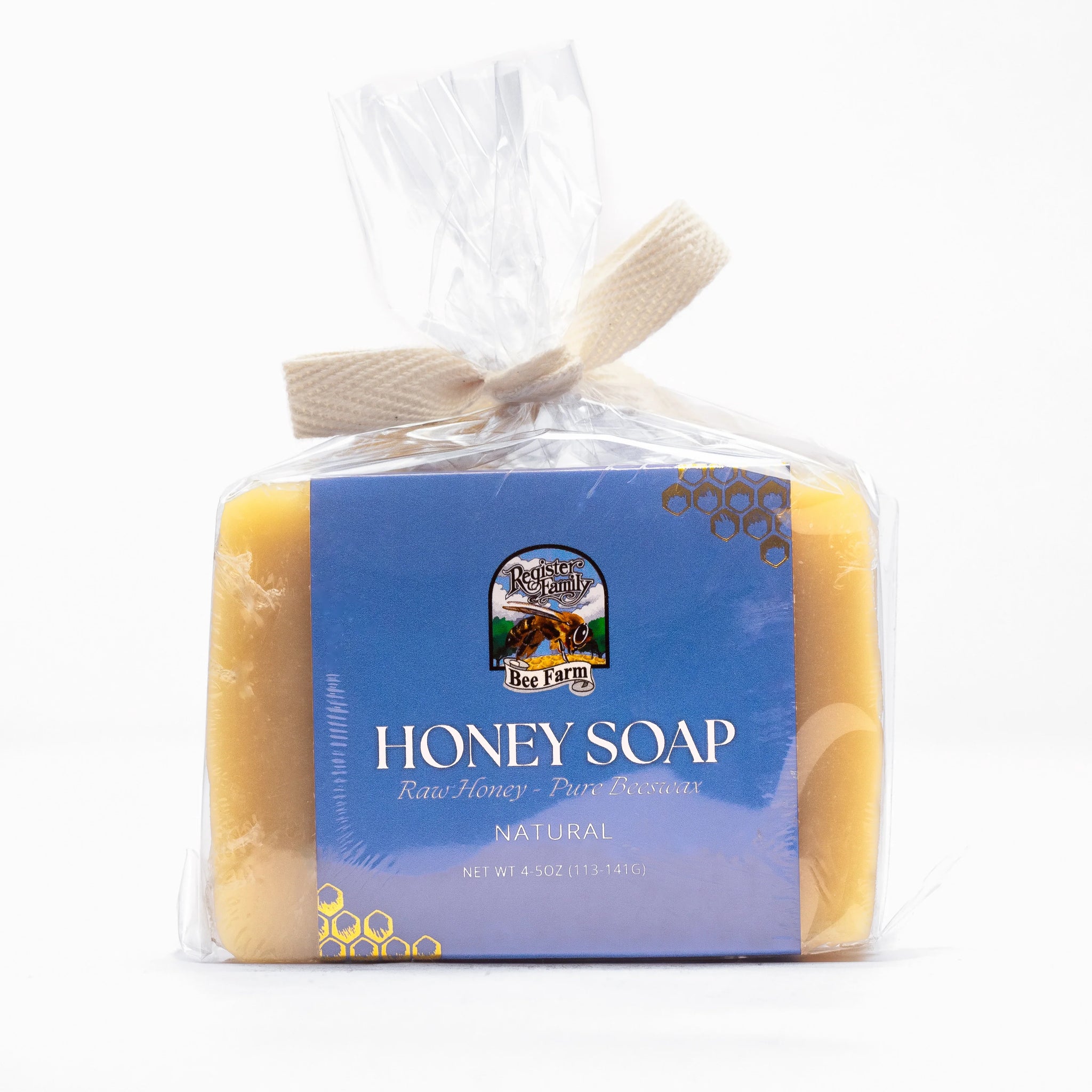 A bar of Register Family Bee Farm honey soap, made with raw honey and pure beeswax, sits wrapped in clear packaging with a woven ribbon tie.

