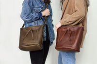 Two women showcase leather tote bags with pockets, wearing denim and corduroy jackets.
