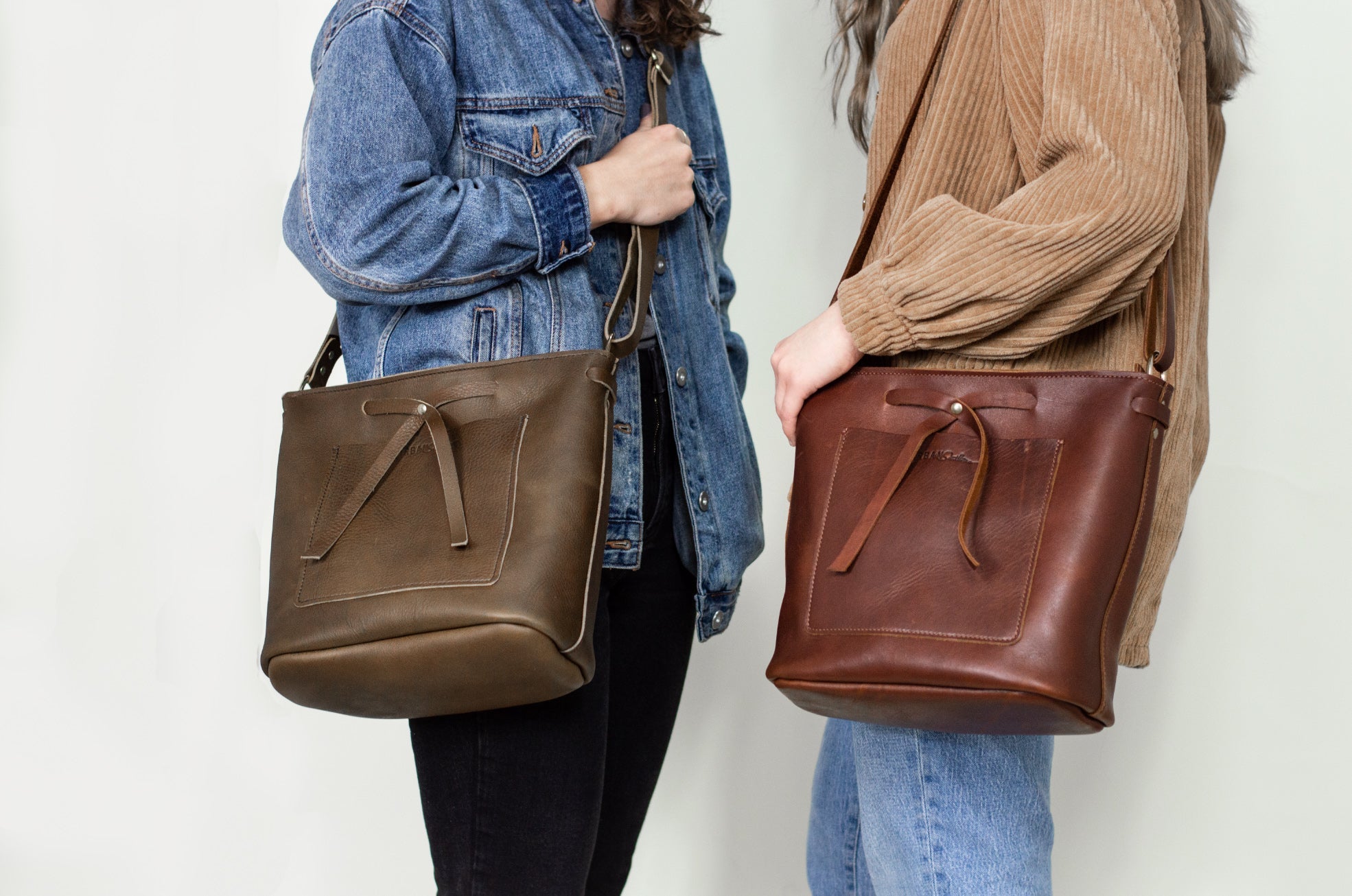 Two women showcase leather tote bags with pockets, wearing denim and corduroy jackets.
