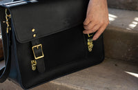 A person fastens the brass buckle on a black leather Urban Southern briefcase.
