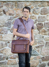 A man carries a brown leather messenger bag with gold buckles, standing against a stone wall.
