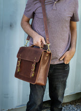 A man models a brown leather satchel bag with brass buckles and a shoulder strap.
