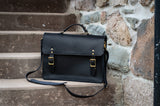 A stylish black leather satchel with gold buckles rests on stone steps against a textured stone wall.
