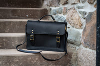 A stylish black leather satchel with gold buckles rests on stone steps against a textured stone wall.
