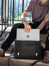 Man places a Microsoft Surface Pro laptop into a black leather satchel while sitting on outdoor steps.
