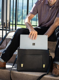 Man places a Microsoft Surface Pro laptop into a black leather satchel while sitting on outdoor steps.
