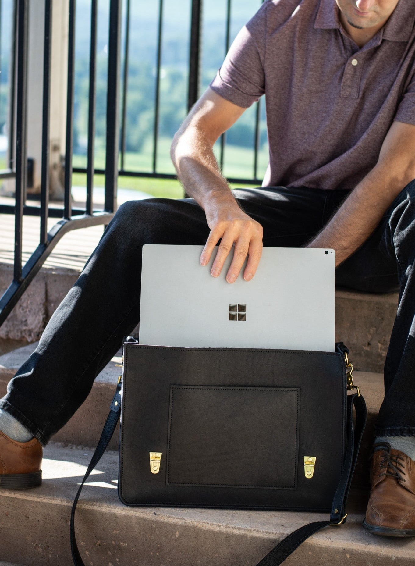 Man places a Microsoft Surface Pro laptop into a black leather satchel while sitting on outdoor steps.
