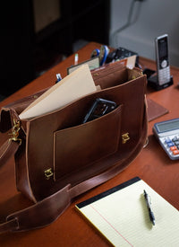 A brown leather satchel, filled with a file and smartphone, sits on a desk next to a notepad and pen, illustrating business organization.
