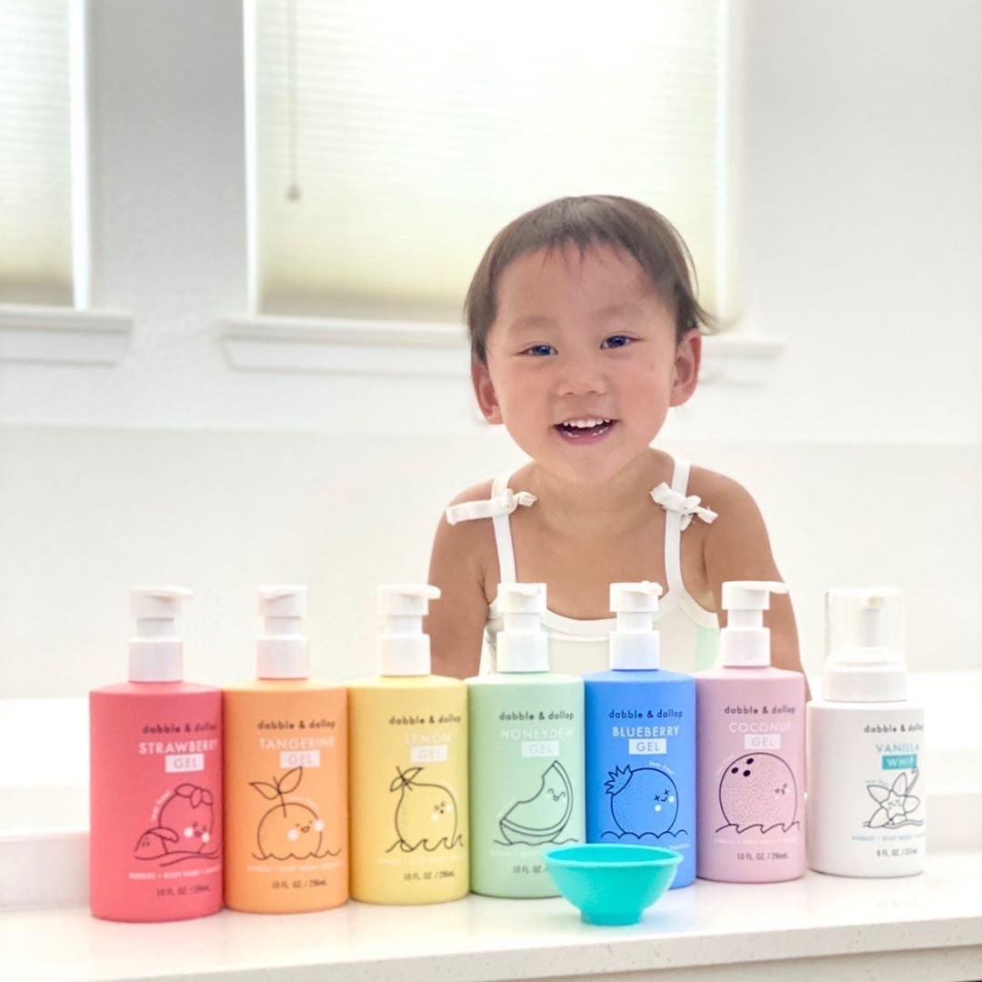 A smiling toddler showcases a colorful row of Dabble & Dollop bath and body gel and whip products in strawberry, tangerine, lemon, honeydew, blueberry, coconut, and vanilla scents.