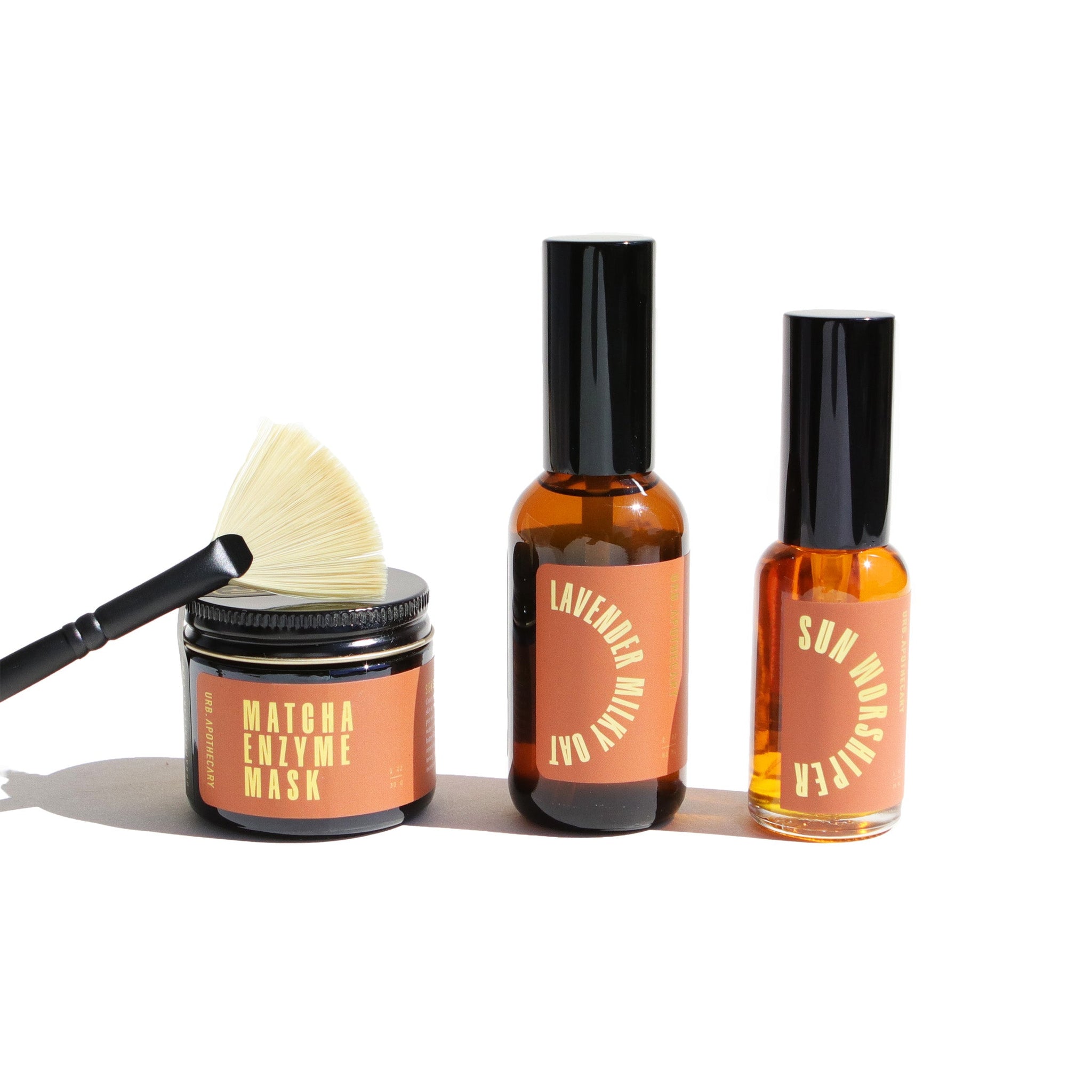 A fan brush rests on a jar of matcha enzyme face mask, next to bottles of lavender milky oat cleanser and sun worshipper oil from URB Apothecary.
