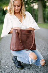 A blonde woman in ripped jeans showcases a brown leather and suede handbag outdoors.
