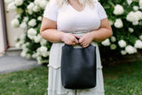 Woman holds a black leather Urban Outfitters tote bag while wearing a white tee and sage green dress outdoors.
