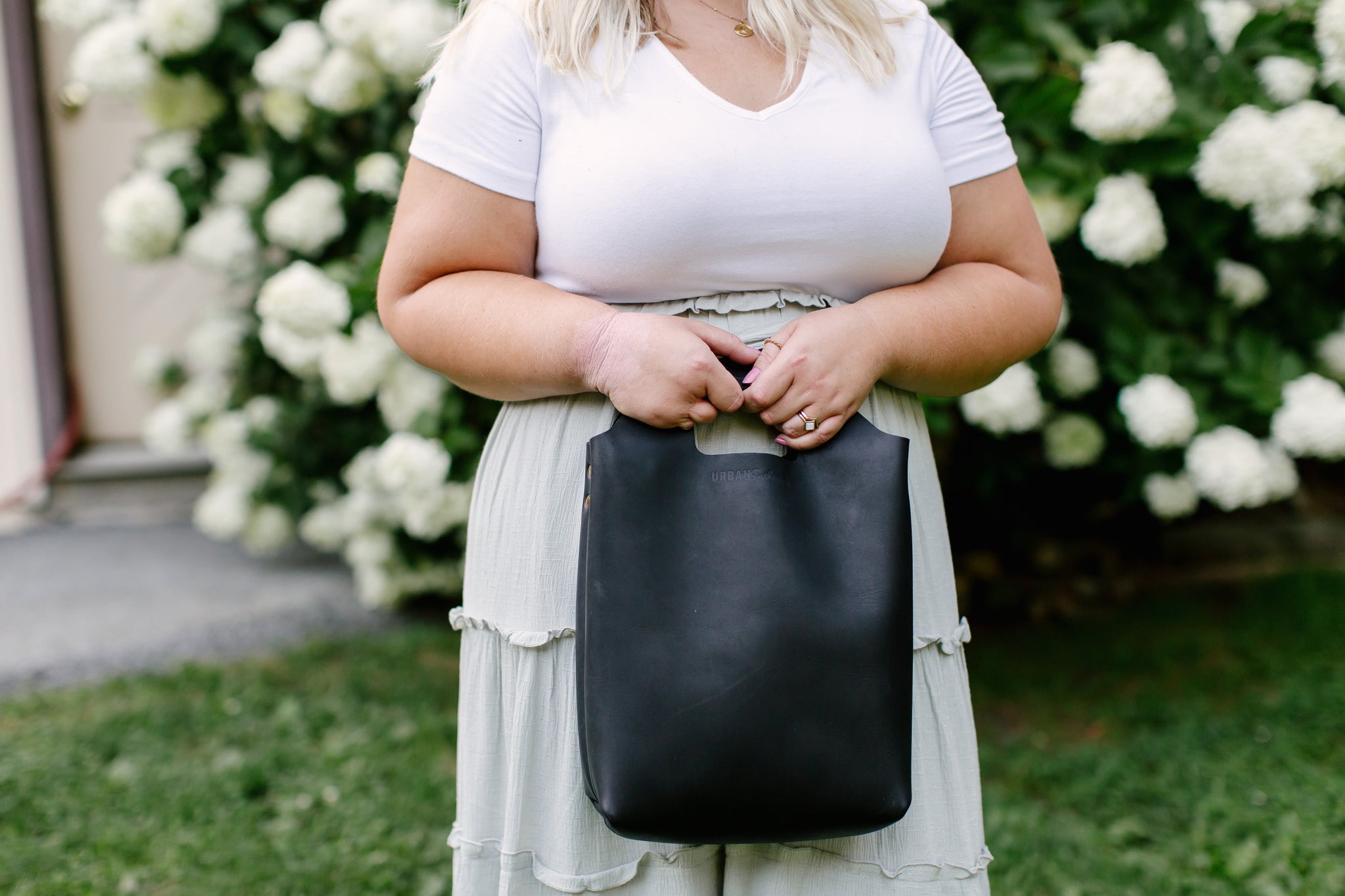 Woman holds a black leather Urban Outfitters tote bag while wearing a white tee and sage green dress outdoors.
