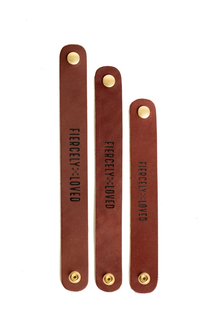 Three brown leather cable straps, each with a brass snap, display the phrase "FIERCELY LOVED".
