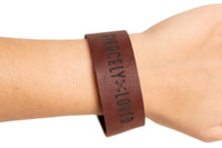 A person wears a brown leather bracelet with the words "FIERCELY LOVED" etched in black.
