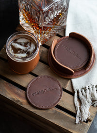 Luxury leather coasters from Urban Southern protect a wooden table holding a whiskey glass and a drink with ice.
