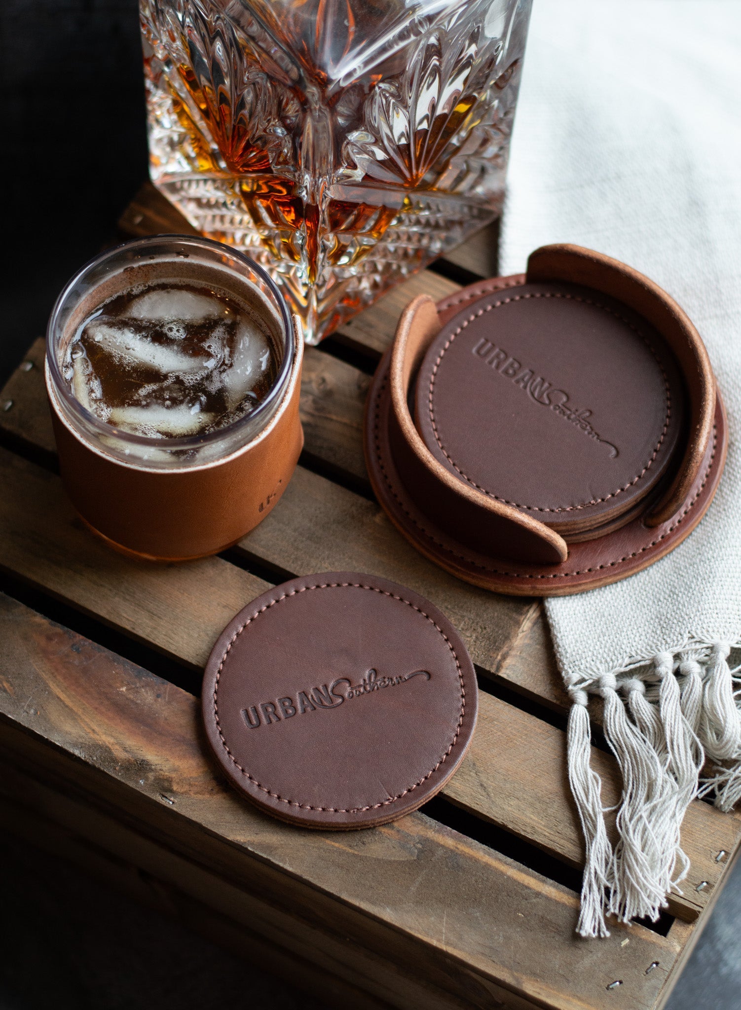 Luxury leather coasters from Urban Southern protect a wooden table holding a whiskey glass and a drink with ice.

