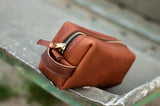 Close-up shows a handcrafted leather dopp kit bag with a gold zipper resting on a weathered metal surface.
