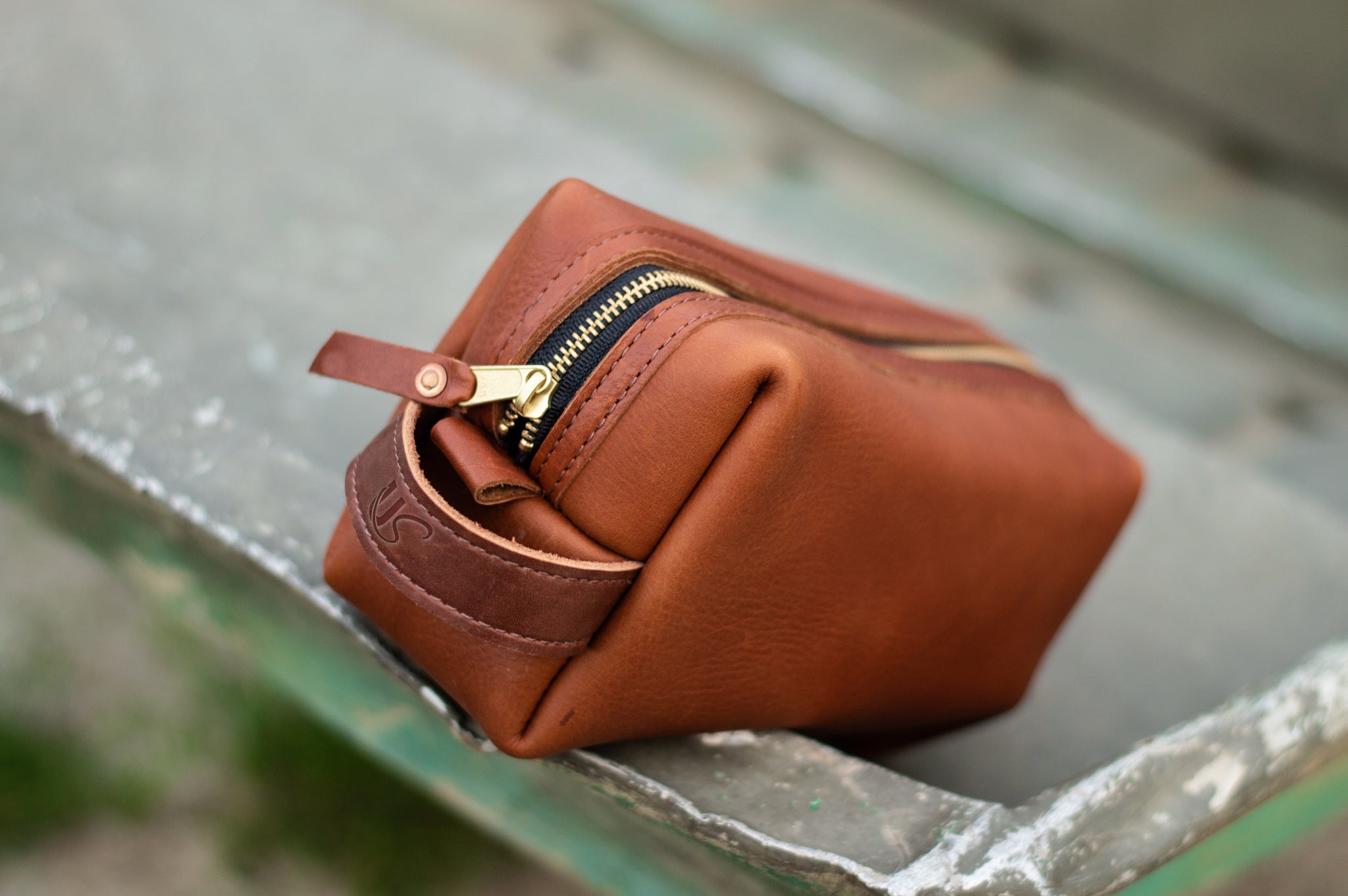 Close-up shows a handcrafted leather dopp kit bag with a gold zipper resting on a weathered metal surface.
