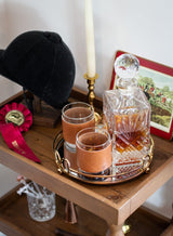 A crystal whiskey decanter and leather-sleeved glasses sit on a gold tray beside a riding helmet and horse show ribbon, evoking an equestrian lifestyle.
