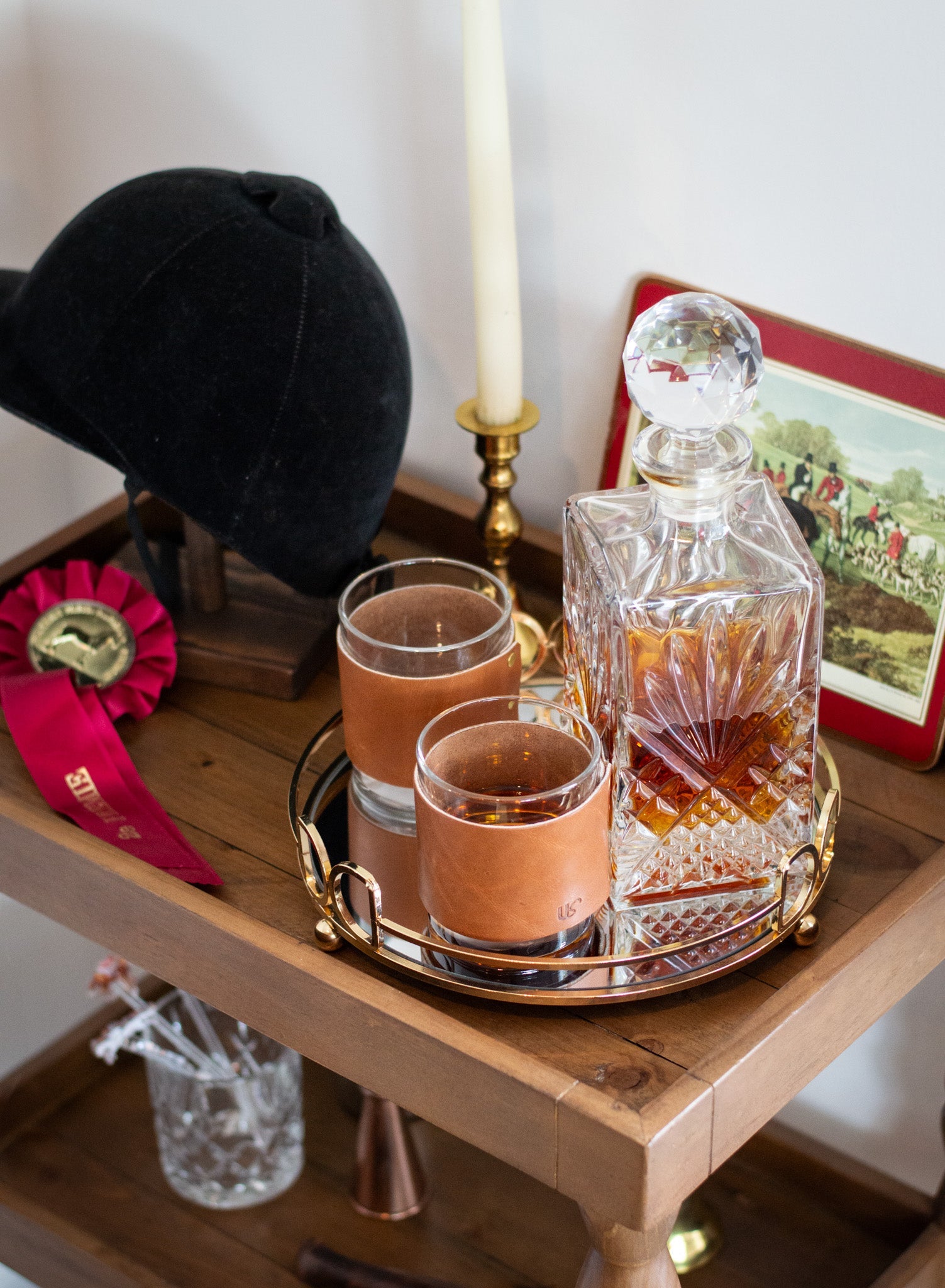 A crystal whiskey decanter and leather-sleeved glasses sit on a gold tray beside a riding helmet and horse show ribbon, evoking an equestrian lifestyle.
