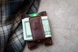 Leather money clip secures folded twenty-dollar bills and a credit card near a plaid jacket.
